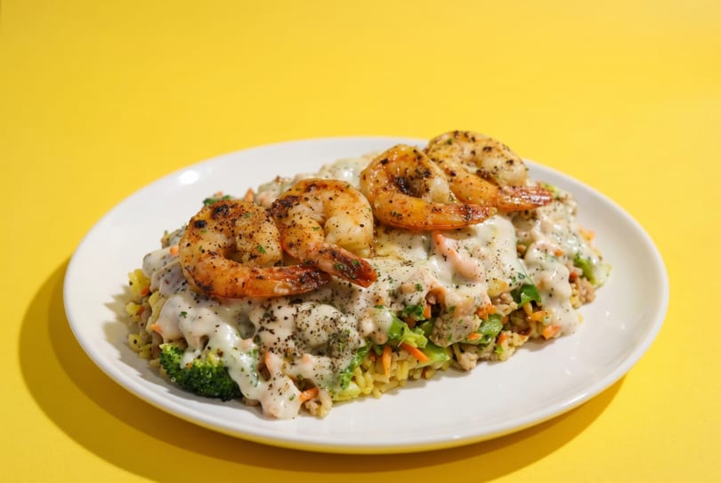 A white plate holds seasoned shrimp over rice, broccoli, and carrots, all covered in a creamy sauce.