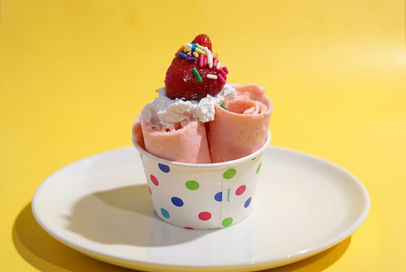 Strawberry rolled ice cream with whipped cream, sprinkles, and a whole strawberry on a white plate.