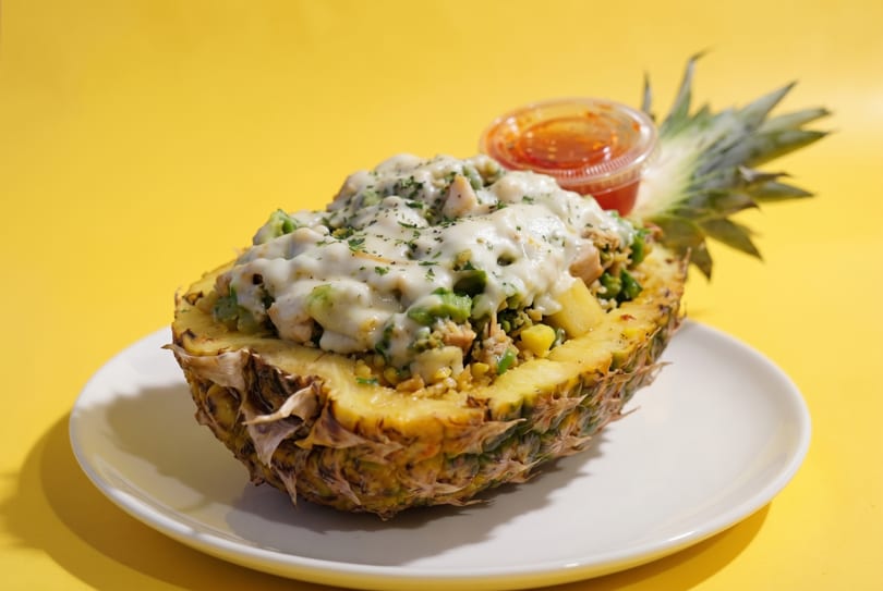 Pineapple boat filled with rice, chicken, vegetables, melted cheese, and a side of sauce.